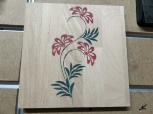 Holzplatte mit gefrästem Blumenmotiv und farbig ausgefüllten Blüten, Blättern und Stängeln.