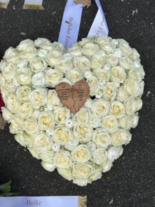 Weißes herzförmiges Trauergesteck mit einem individuell gravierten Holzherz in der Mitte.